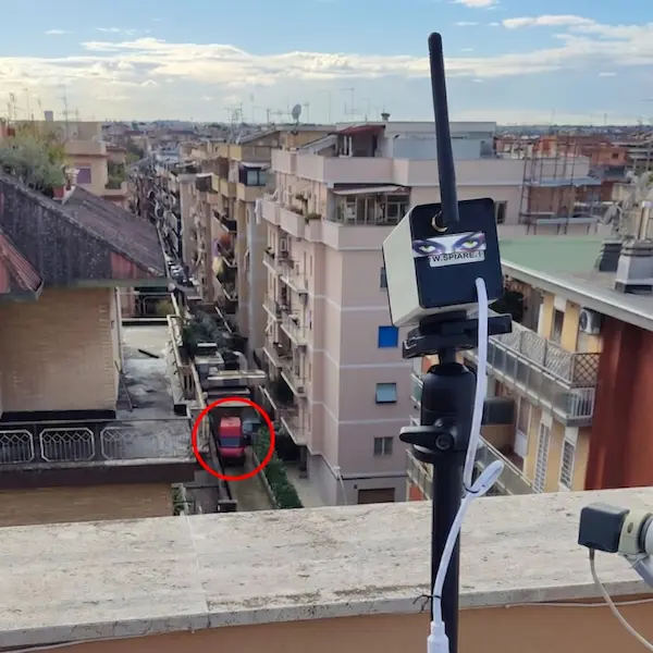 Telecamera IP WiFi spia con zoom ottico 36x posizionata su treppiede su terrazzo con vista strada a Roma, cerchio rosso indica il soggetto monitorato a distanza